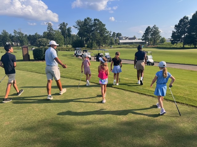 A coach leading a group of junior golfers onto the course for their 9-hole event